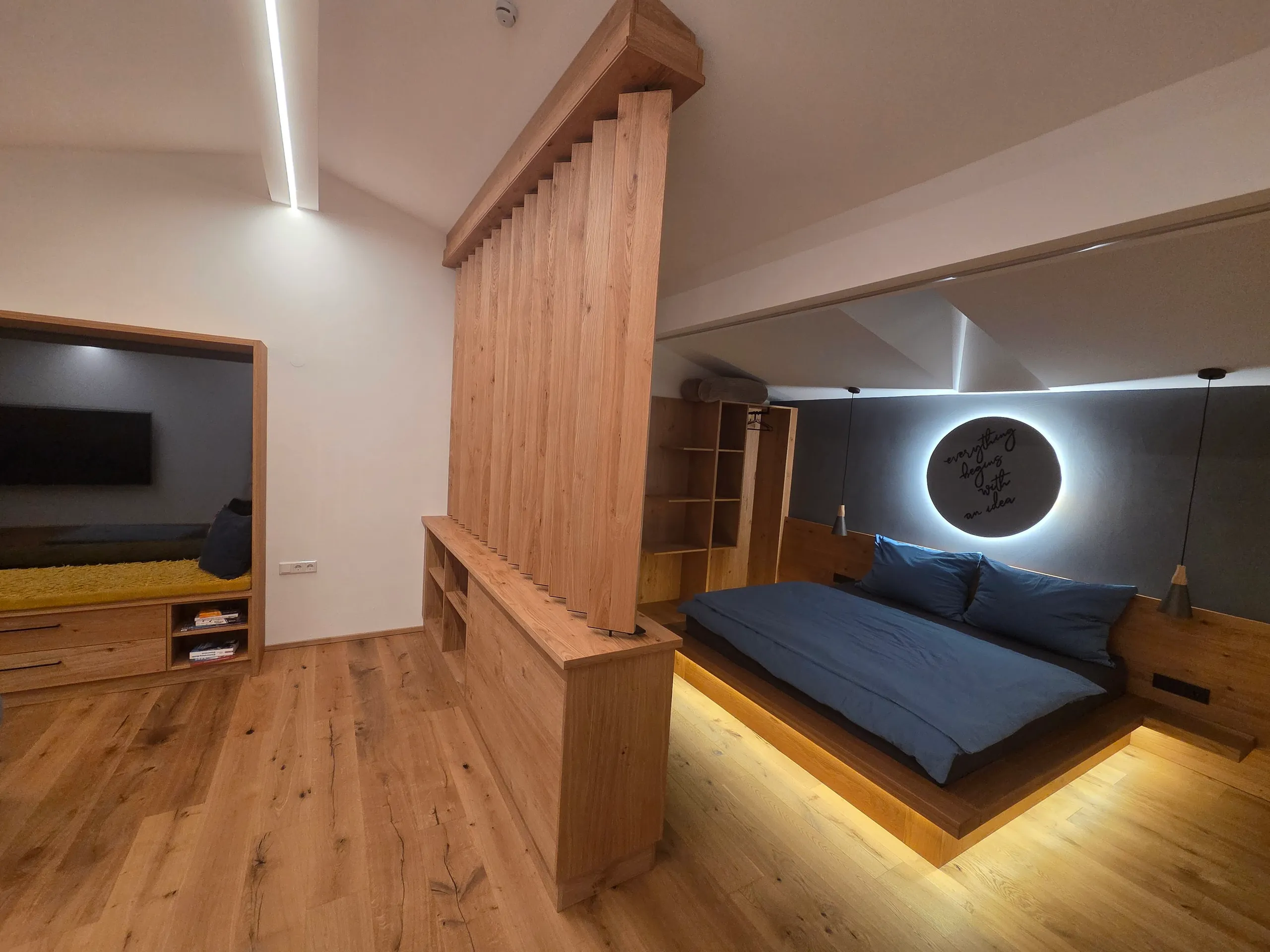 Blick über den Lamellen-Raumteiler auf das beleuchtete Kingsize-Bett, offenes Kleiderregal und Fernsehwand