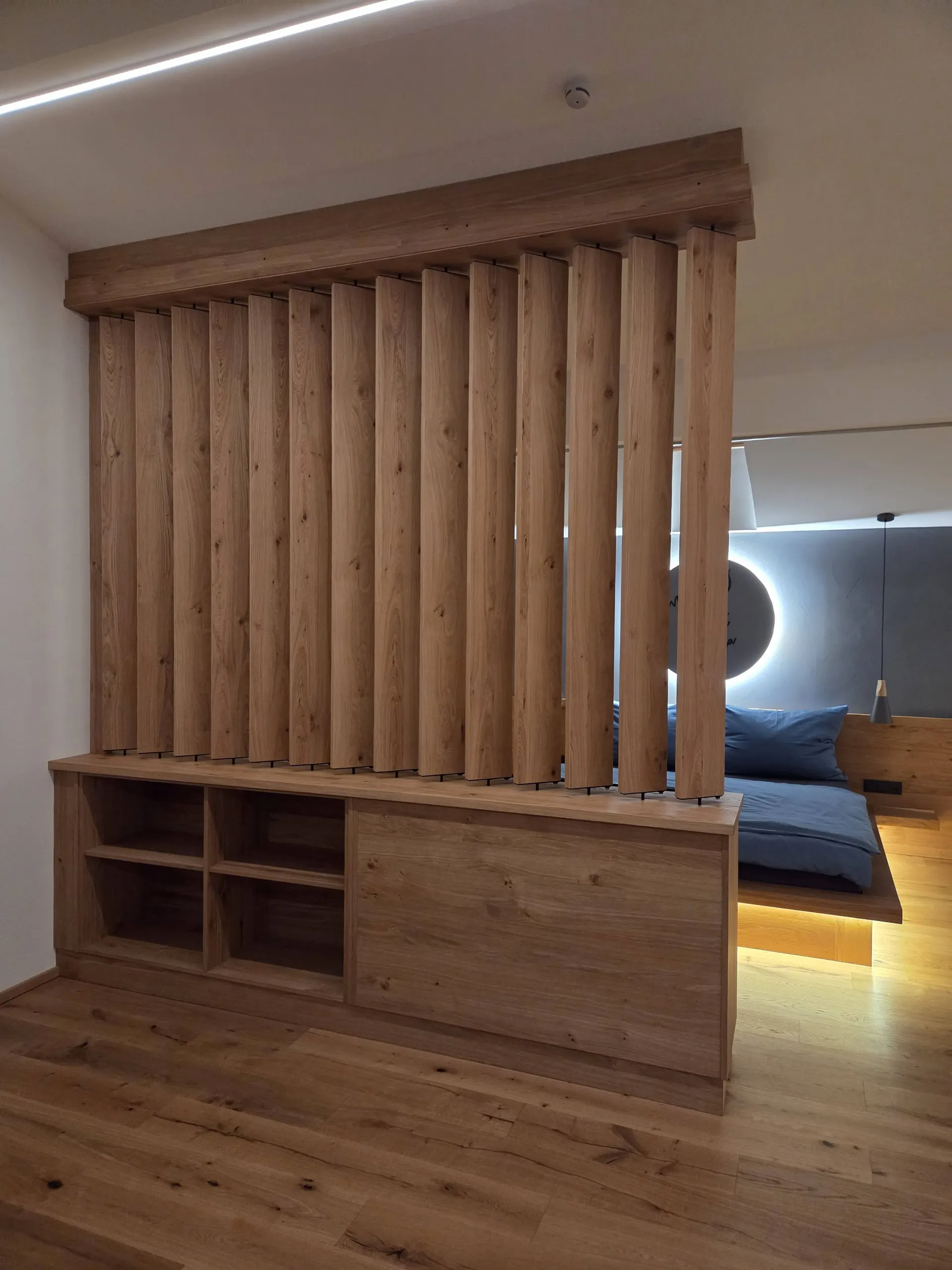 Gedrehter Lamellen-Raumteiler über dem Sideboard – vom Wohnbereich fällt der Blick durch auf das Bett